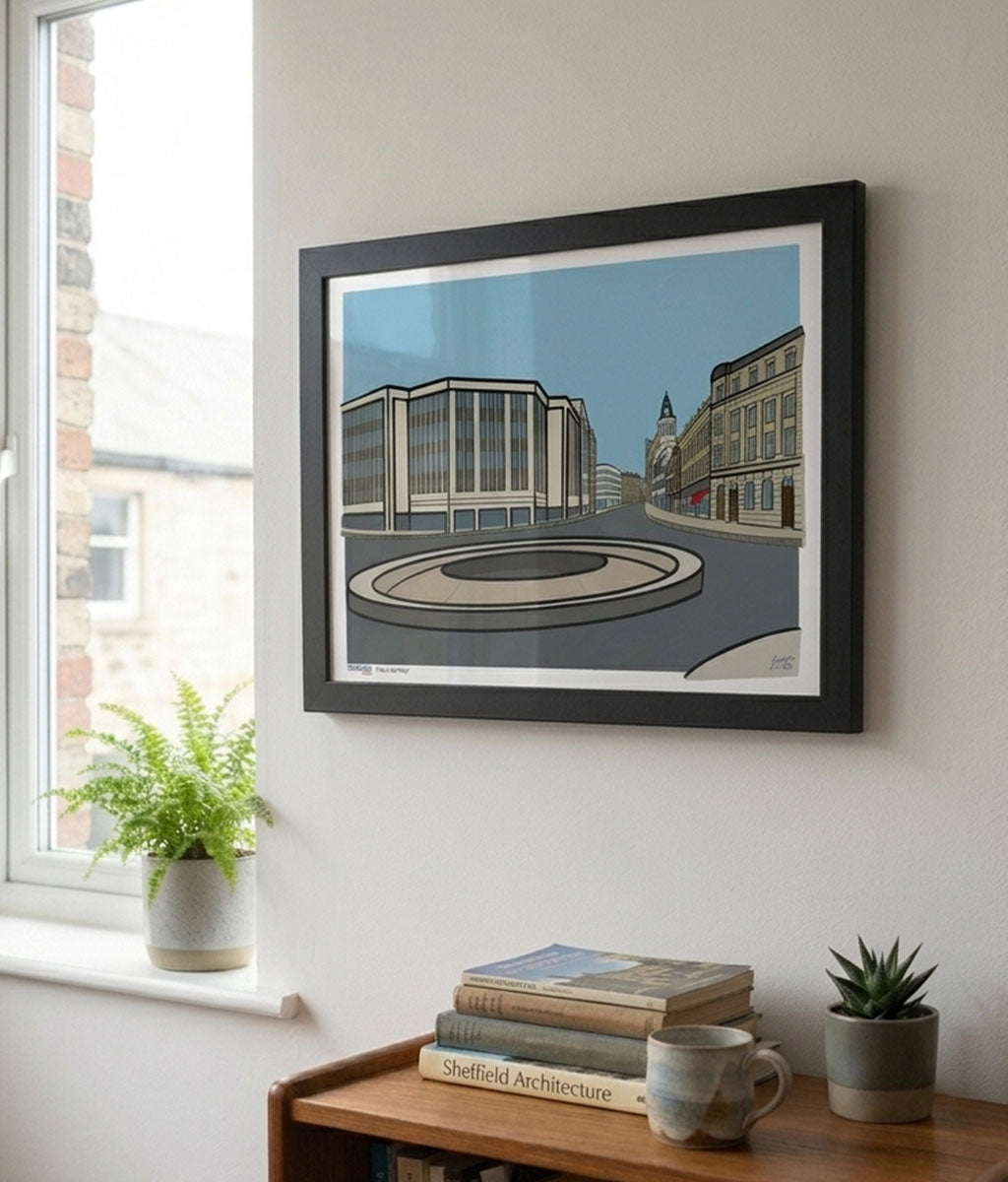 Framed artwork of a cityscape on a wall in a room with a window and books.
