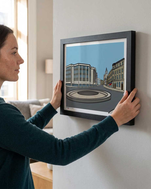 A woman in a dark teal sweater holds a black-framed art print of the Hole in the Road in Sheffield against a light gray wall in a modern living room setting. The print is a stylized illustration of a street scene with buildings and a roundabout.