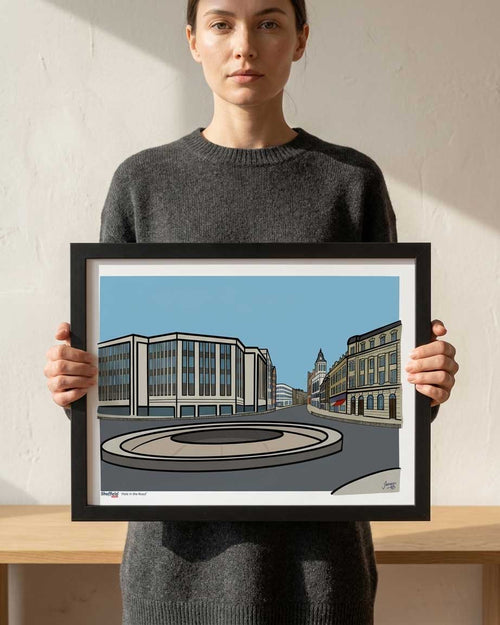 A woman in a dark gray sweater holds a framed art print depicting The Hole in the Road in Sheffield, a modernist street scene with a roundabout and buildings under a blue sky.
