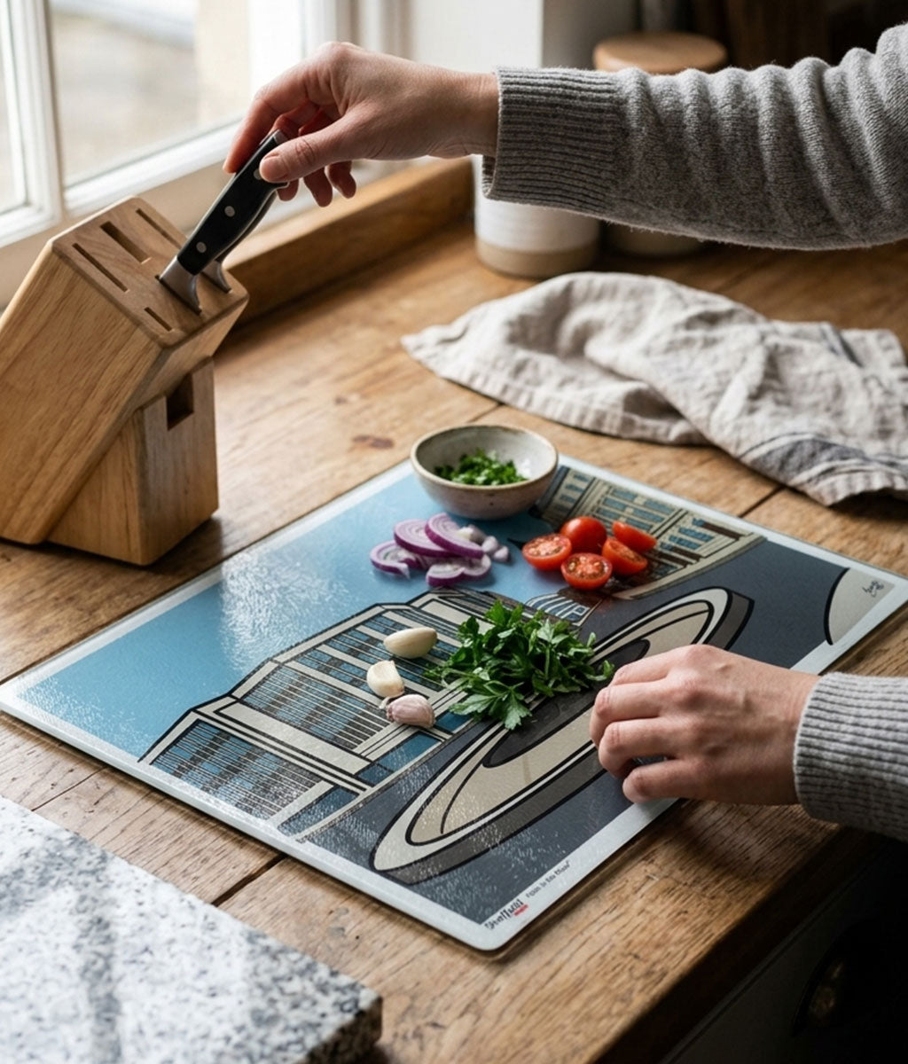 Person preparing food on a glass cutting board with a knife block and ingredients on a wooden table.