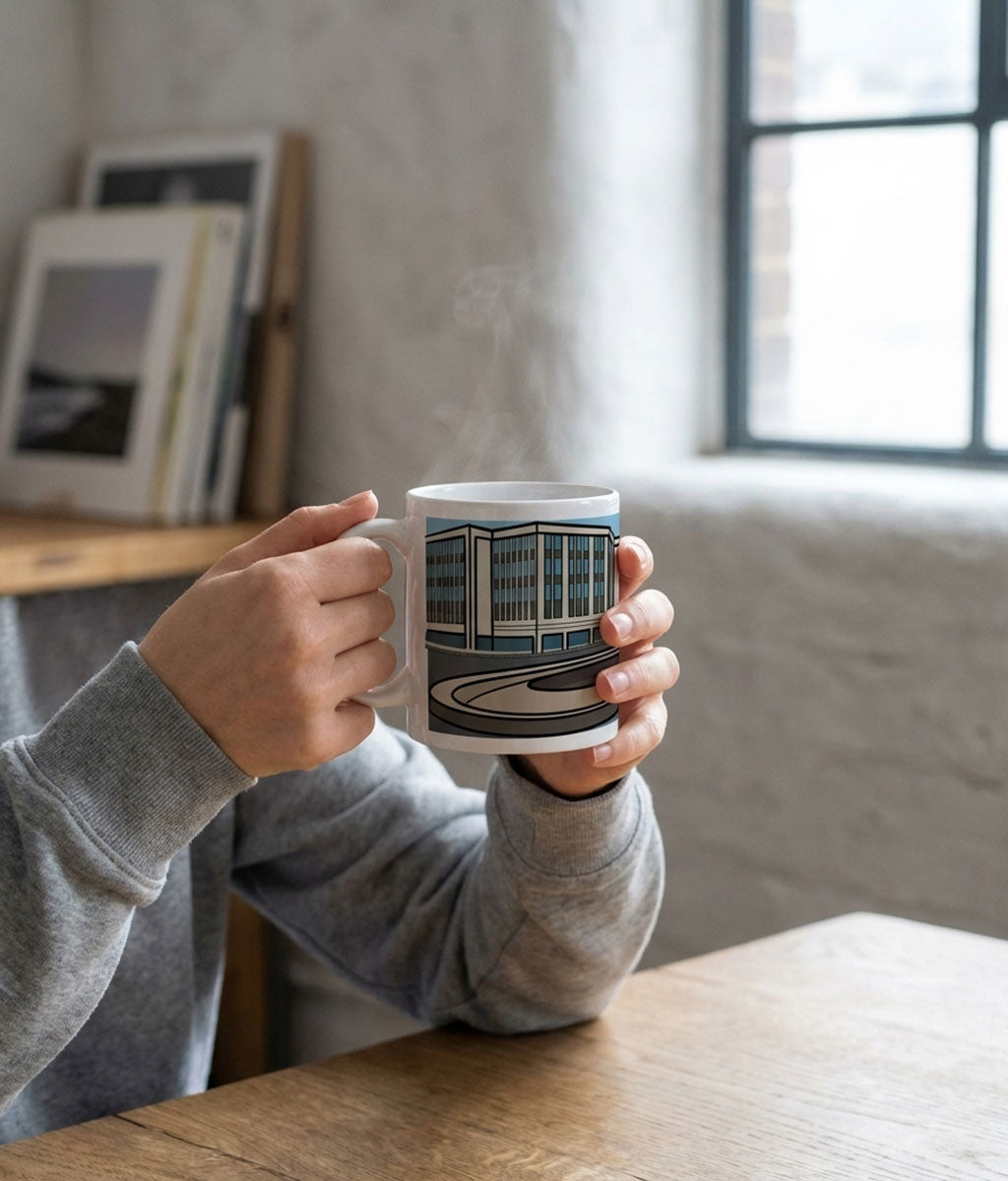 Person holding a mug with a building design, sitting at a table by a window.
