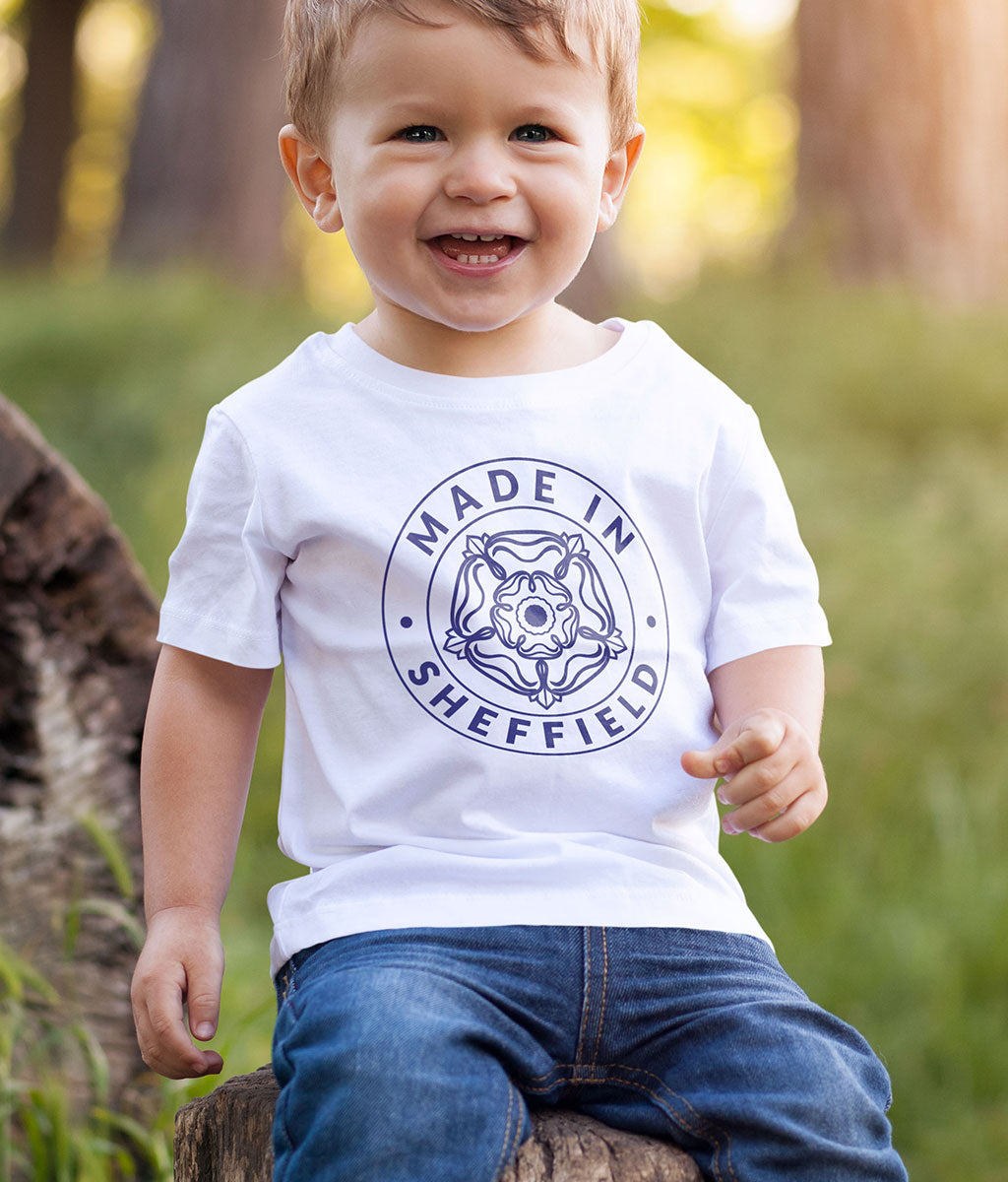 Young child wearing a white Made in Sheffield baby t-shirt with the exclusive Yorkshire Rose badge design in navy, seated outdoors.
