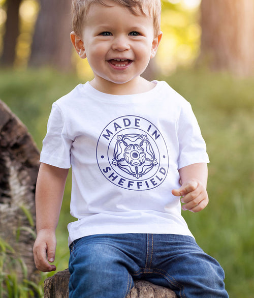 Young child wearing a white Made in Sheffield baby t-shirt with the exclusive Yorkshire Rose badge design in navy, seated outdoors.
