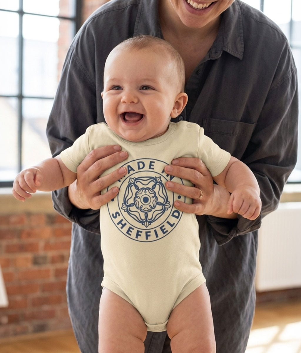 Baby wearing a 'Made in Sheffield' onesie being held by an adult indoors.