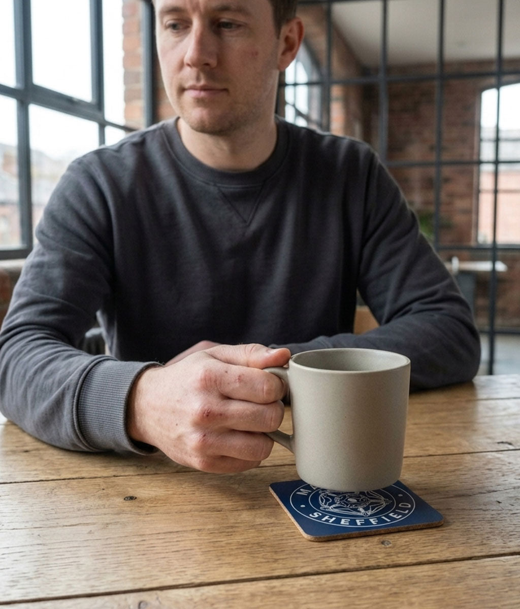 Man holding a mug on a wooden table with a window and brick wall background, blue 'Made in Sheffield' coaster in forreground