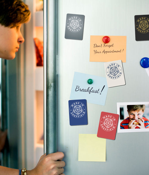 Made in Sheffield ceramic fridge magnets in five colourways — Steel, Royal Blue, Cream, Navy and Red — displayed on a fridge door alongside notes and photos.