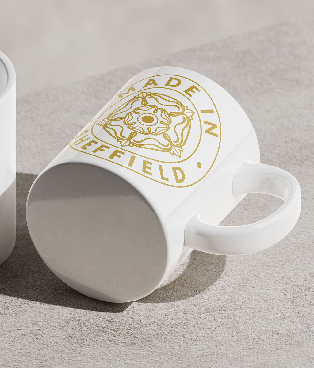 White mug with gold 'Made in Sheffield' yorkshire rose design laid on it's side on a grey counter