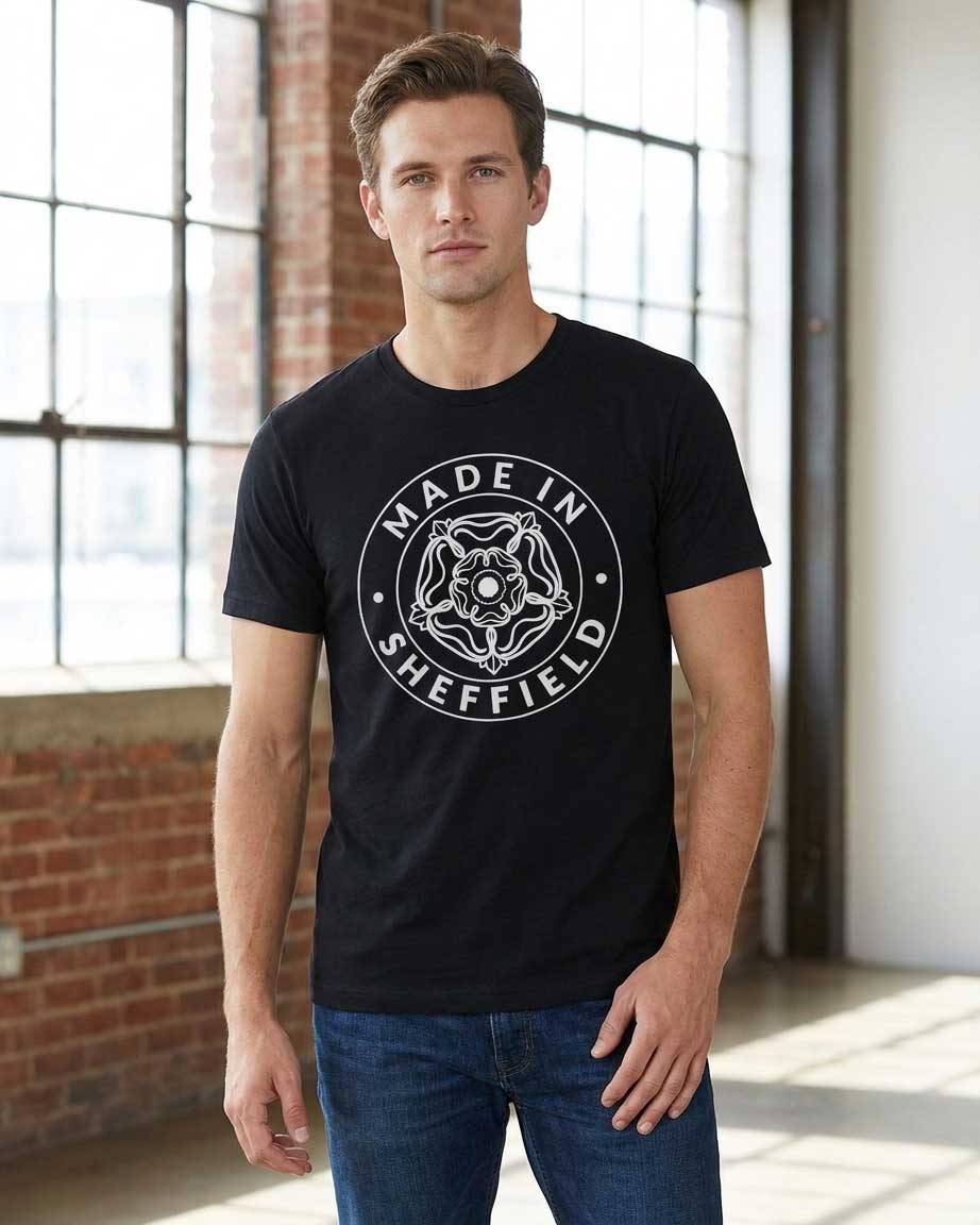 A man wearing a black crewneck t-shirt with a white circular graphic that reads 'MADE IN SHEFFIELD' around a central Yorkshire Rose design, standing in a bright industrial loft with brick columns and large windows.