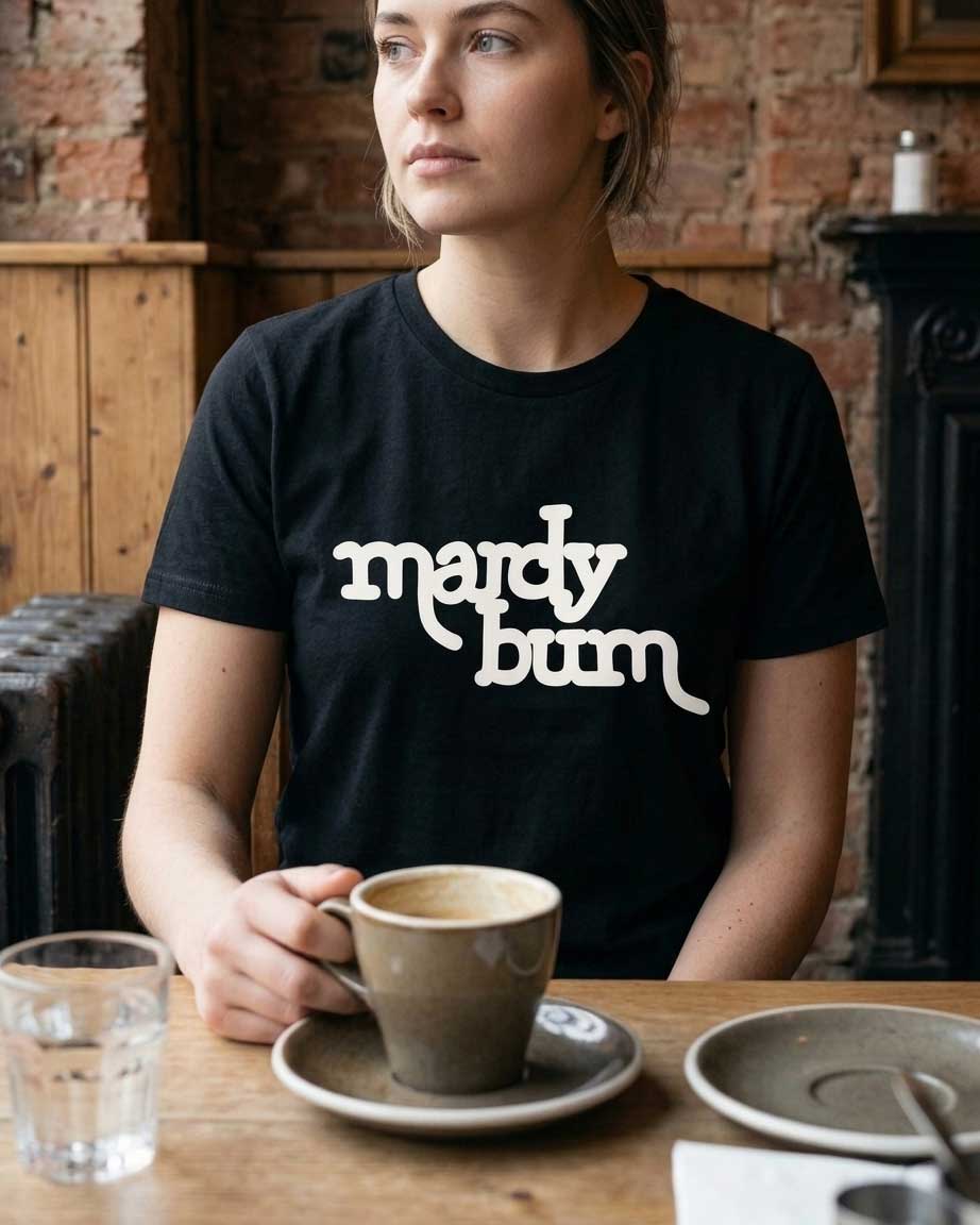 A woman sitting at a wooden table in a rustic cafe setting, wearing a black t-shirt with the phrase 'mardy bum' printed in white stylized text across the chest.