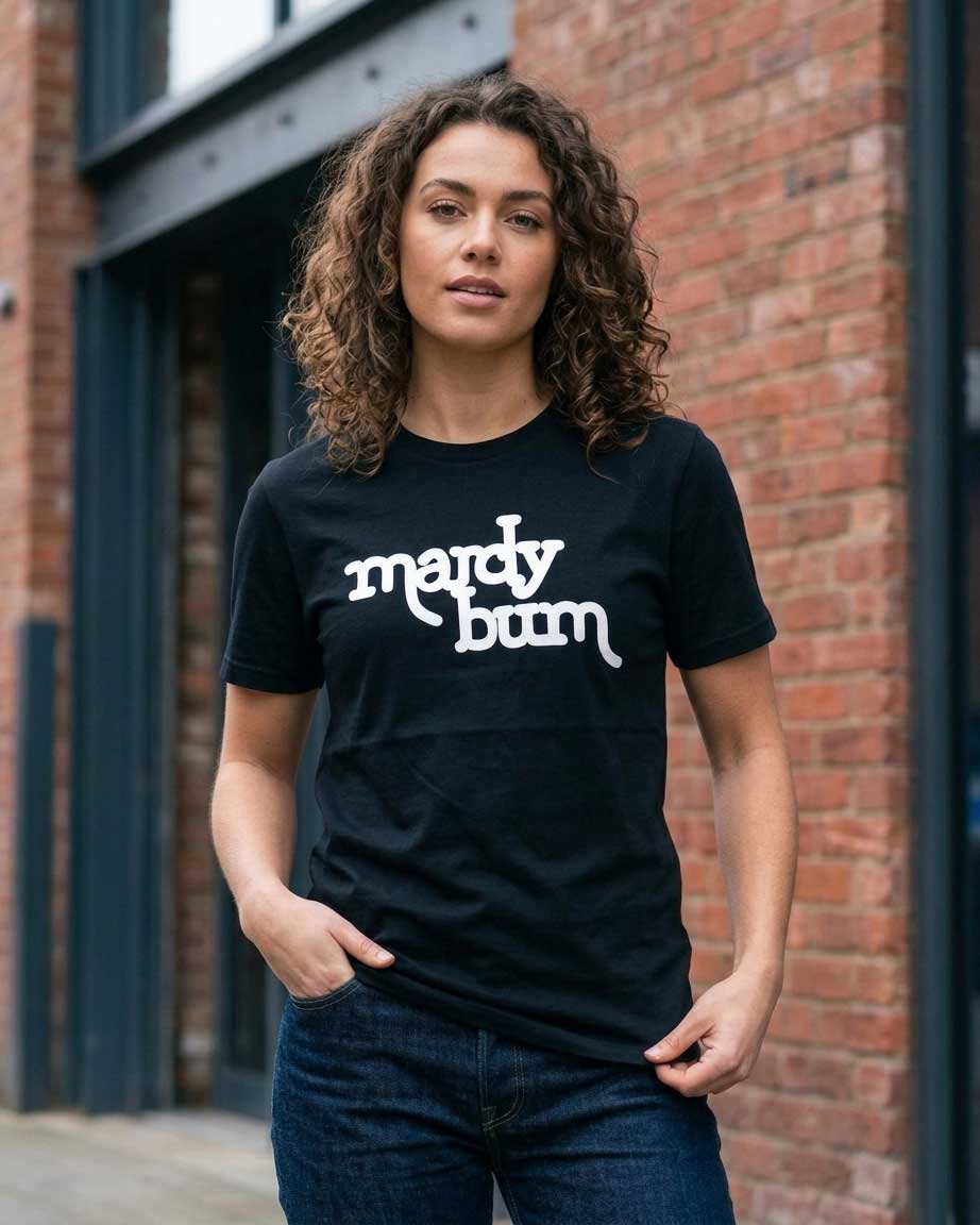 A woman wearing a black crewneck t-shirt with the phrase 'mardy bum' printed in white stylized script across the chest, standing in front of a red brick building.