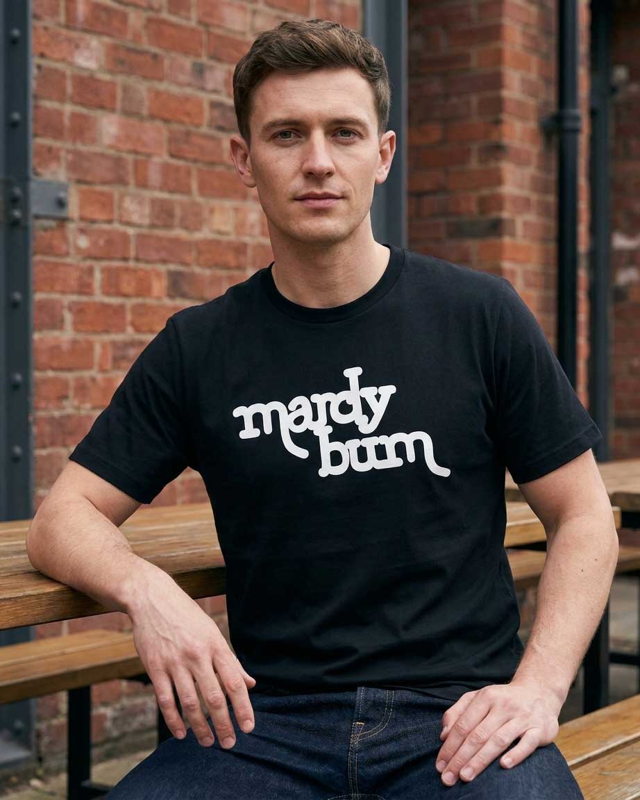 A man wearing a black crew-neck t-shirt with 'mardy bum' printed in white stylized text across the chest, sitting on a wooden bench in front of a red brick wall.