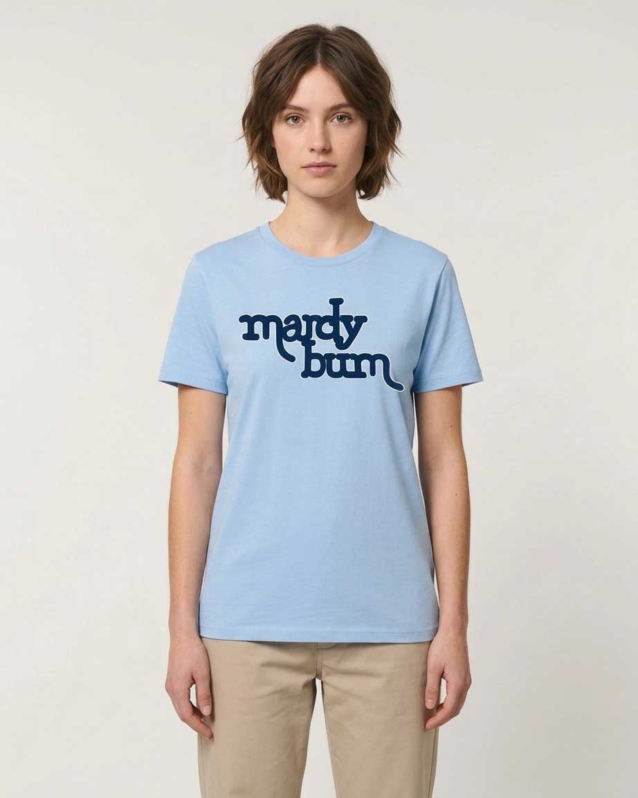 A woman modeling a light blue crewneck t-shirt with 'mardy bum' printed in dark navy blue across the chest, paired with beige trousers, against a plain off-white background.