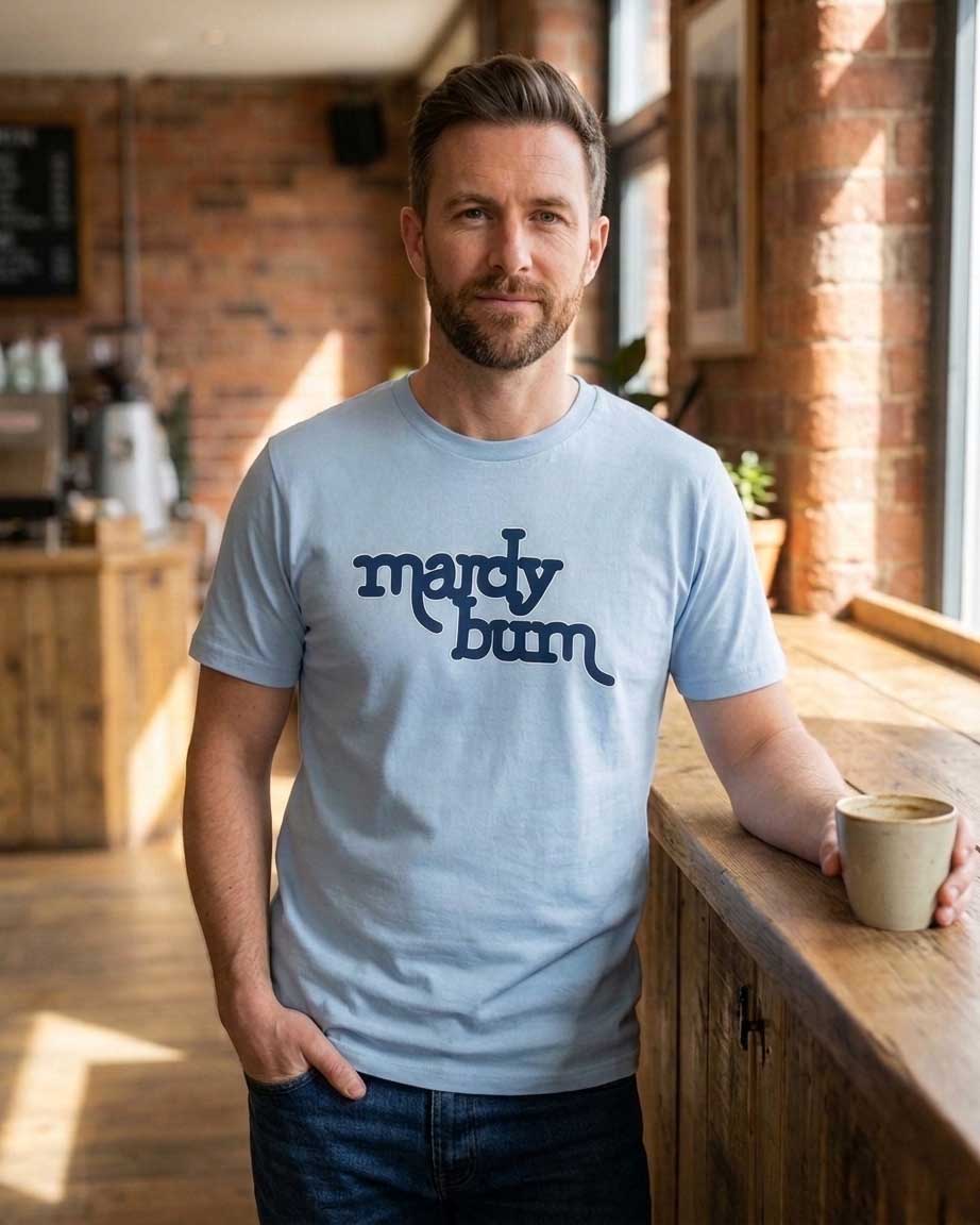 A man wearing a light blue crewneck t-shirt with 'mardy bum' inspired by Arctic Monkeys printed in dark blue script across the chest, standing in a warm, sunlit coffee shop with brick walls and holding a ceramic mug.