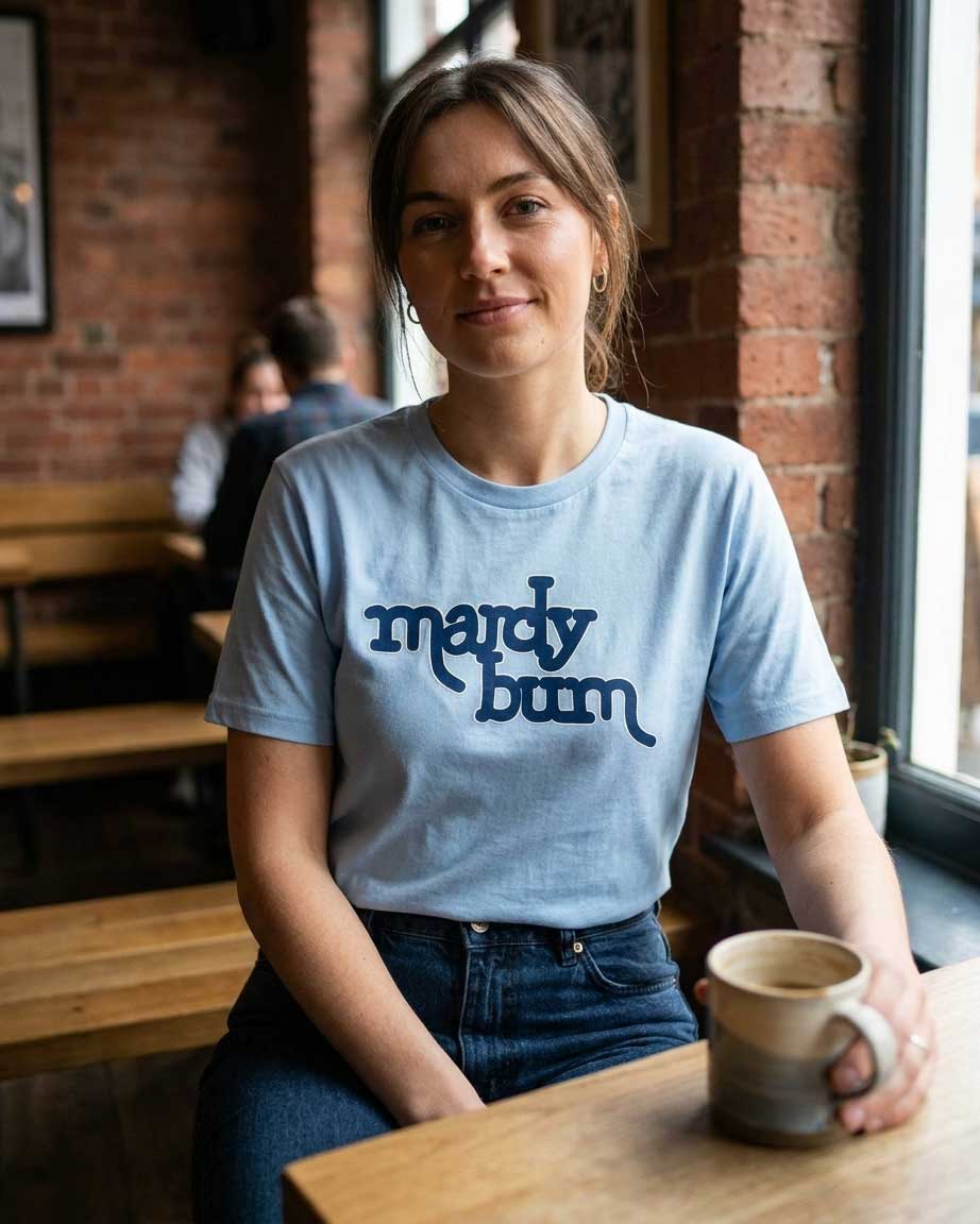 A woman wearing a light blue crewneck t-shirt with 'mardy bum' inspired by Arctic Monkeys printed in dark blue script across the chest, standing in a warm, sunlit coffee shop with brick walls and holding a ceramic mug.
