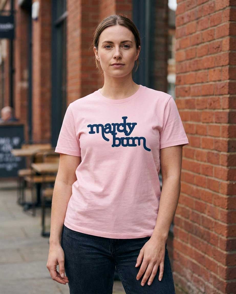 A woman wearing a light pink crewneck t-shirt with the words 'mercy bum' printed in dark blue script across the chest, standing outdoors against a red brick wall.