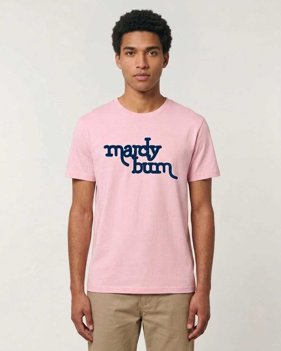 A man wearing a light pink crewneck t-shirt with 'mardy bum' printed in navy blue across the chest, paired with khaki pants, against a light gray studio background.