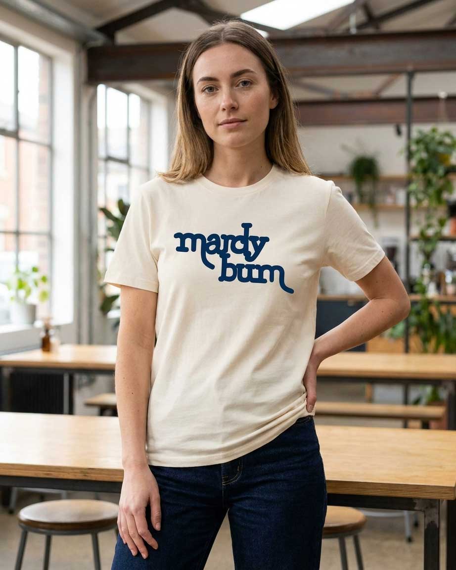 A woman wearing a cream-colored t-shirt with 'mardy bum' printed in dark blue script across the chest, standing in a bright, modern loft-style room with wooden tables and windows.