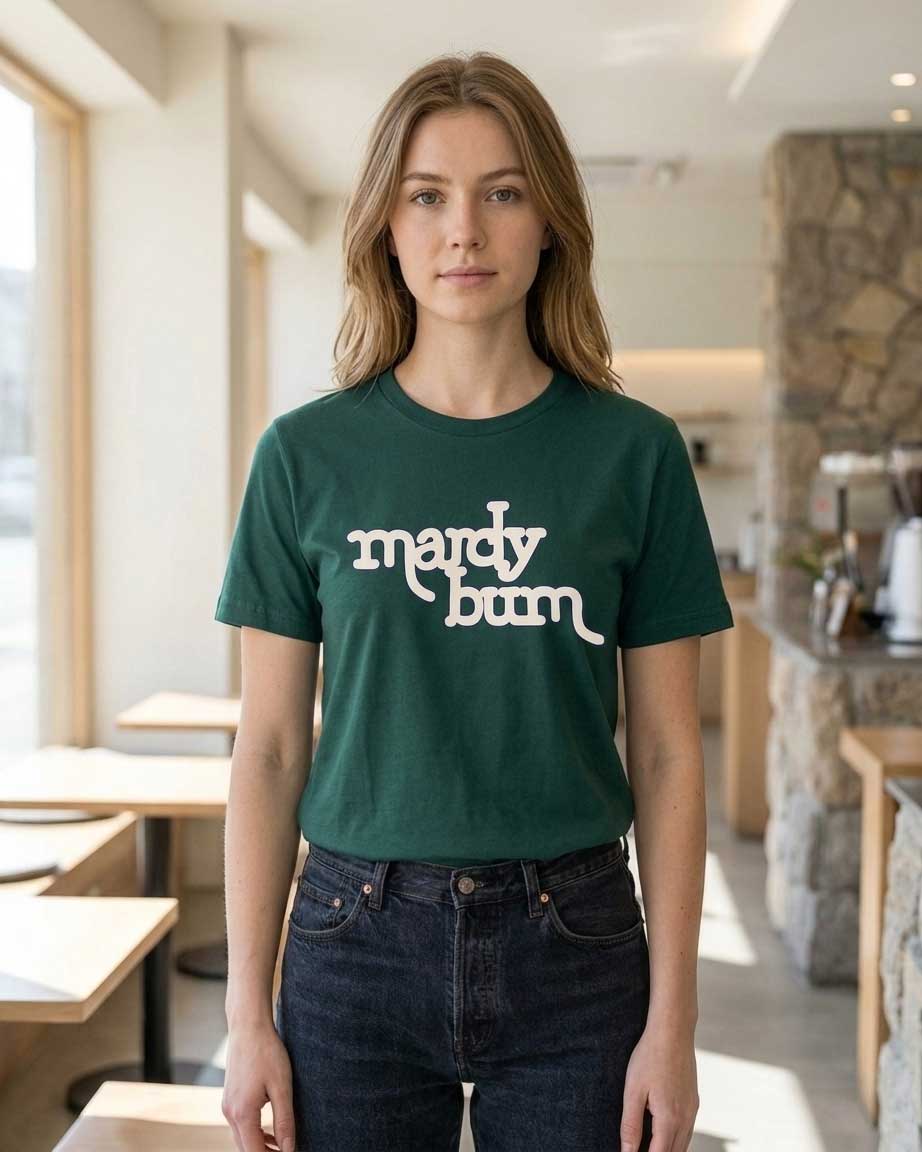 A woman wearing a dark green crewneck t-shirt with the text 'mardy bum' printed in white across the chest, paired with dark blue jeans, standing in a brightly lit cafe interior.