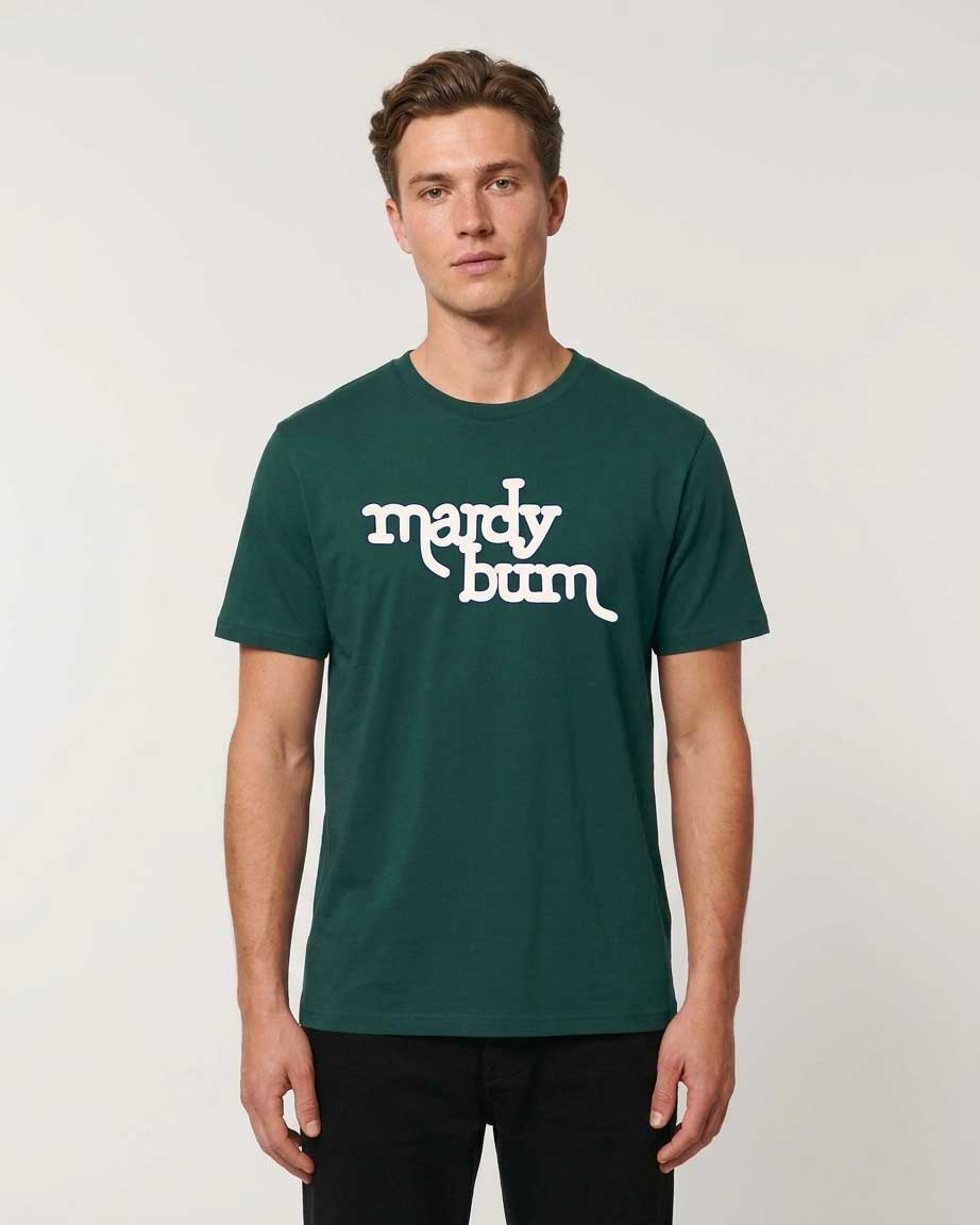 A man wearing a dark green crewneck t-shirt with 'mardy bum' printed in white script across the chest, paired with black pants, against a light gray studio background.