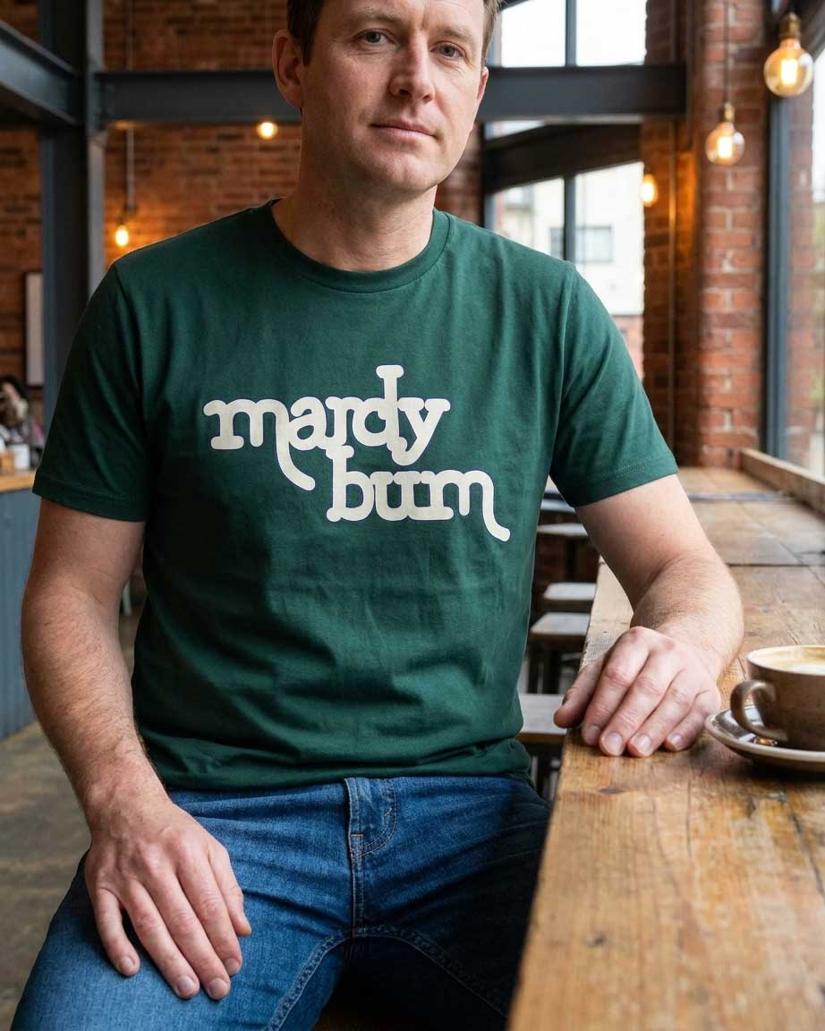 A man wearing a dark green t-shirt with the text 'mardy bum' printed in white across the chest, sitting at a wooden table in a cozy cafe setting.