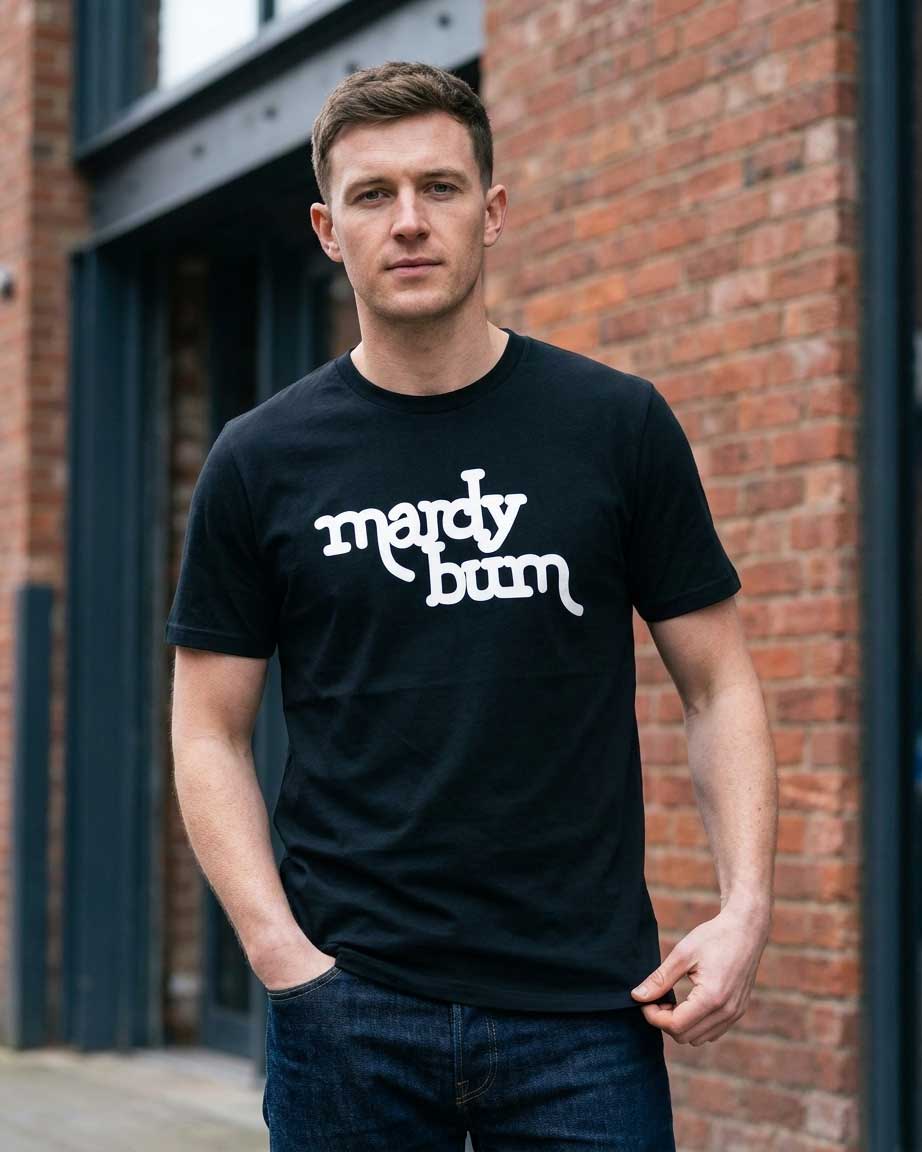 A man wearing a black crewneck t-shirt with the phrase 'mardy bum' printed in white stylized script across the chest, standing in front of a red brick building.