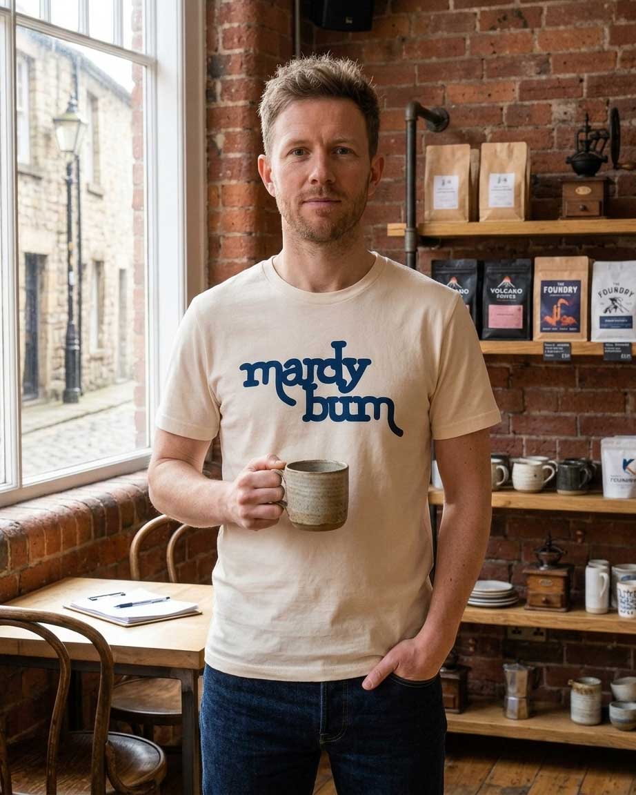 A man wearing a cream-colored t-shirt with 'mardy bum' printed in blue script, holding a ceramic mug inside a rustic brick coffee shop.