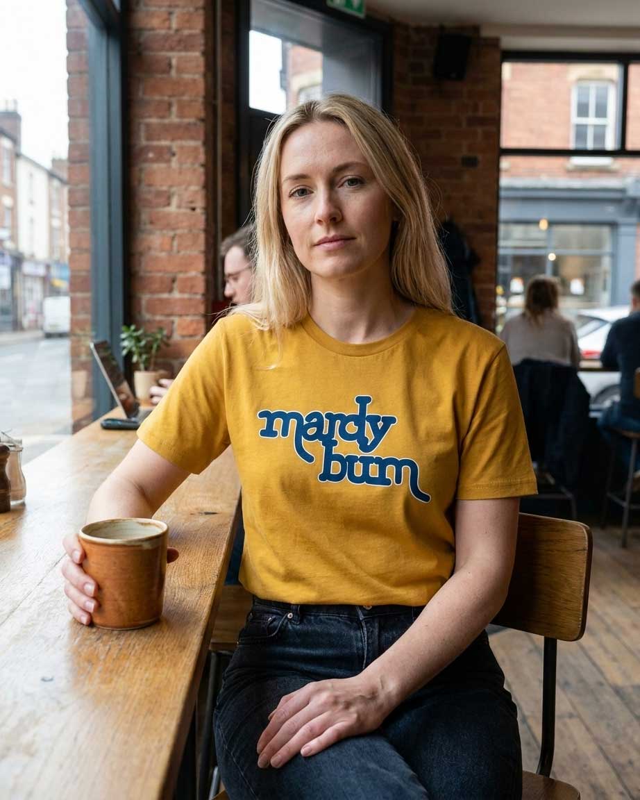 A woman with blonde hair sits at a wooden table in a cafe, wearing a mustard yellow t-shirt with 'mardy bum' printed in blue script. She holds a ceramic mug.
