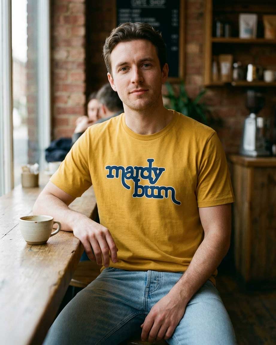 A man sitting at a wooden table in a cozy cafe setting, wearing a mustard yellow t-shirt with 'mardy bum' printed in blue across the chest, paired with light blue jeans.
