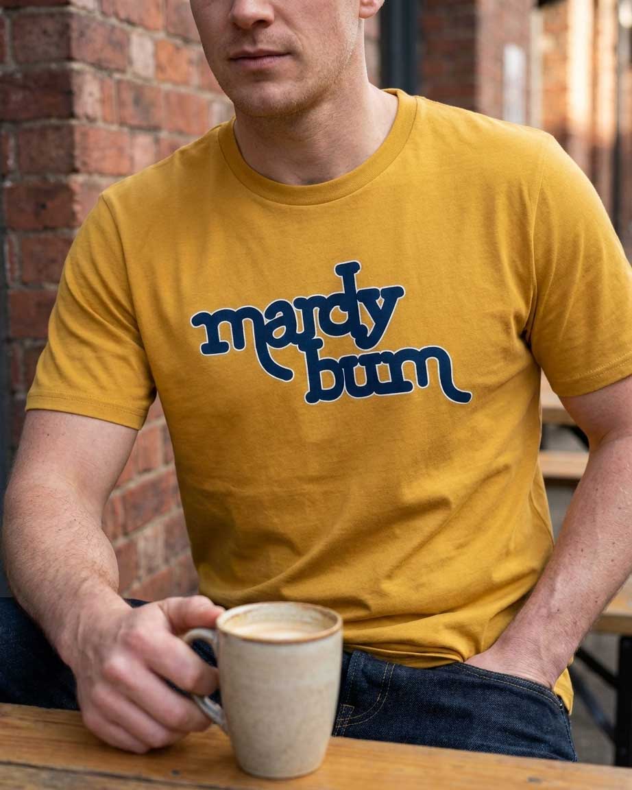 A man wearing a mustard yellow t-shirt with the phrase 'mardy bum' printed in dark blue across the chest, holding a ceramic mug at a wooden table with a brick wall background.