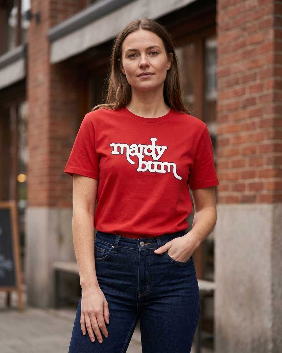 A woman wearing a red crewneck t-shirt with 'mardy bum' printed in white script across the chest, paired with dark blue jeans, standing outdoors in front of a brick building.