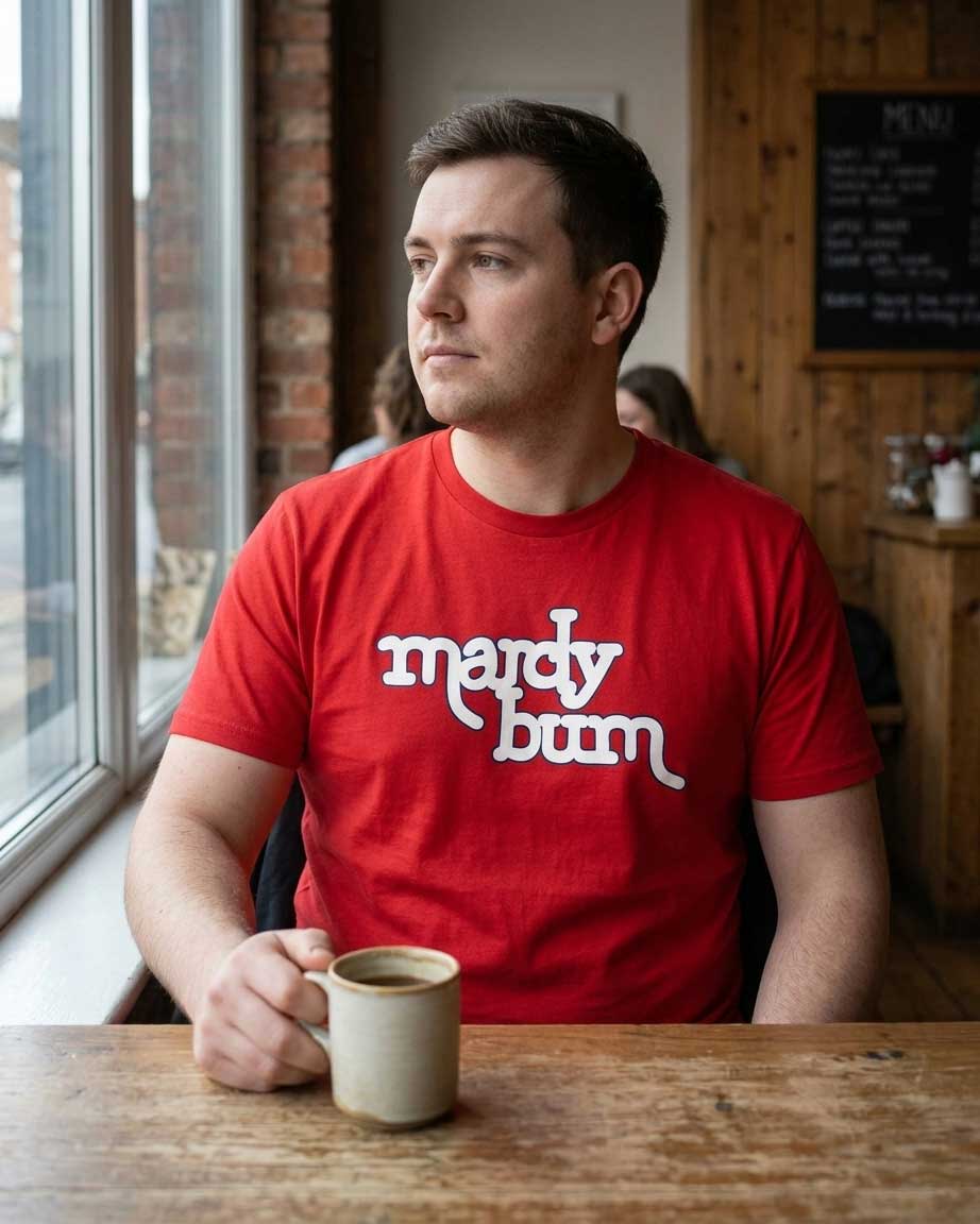 A man wearing a red t-shirt with the white text 'mardy bum' sits at a wooden table in a cafe, holding a ceramic mug.