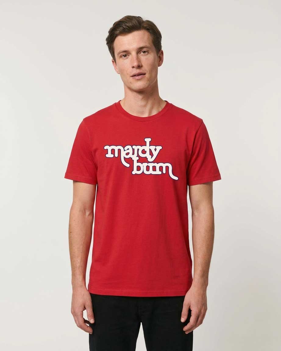 A man wearing a red crewneck t-shirt with 'mardy bum' printed in white script across the chest, paired with black pants, standing against a light gray studio background.