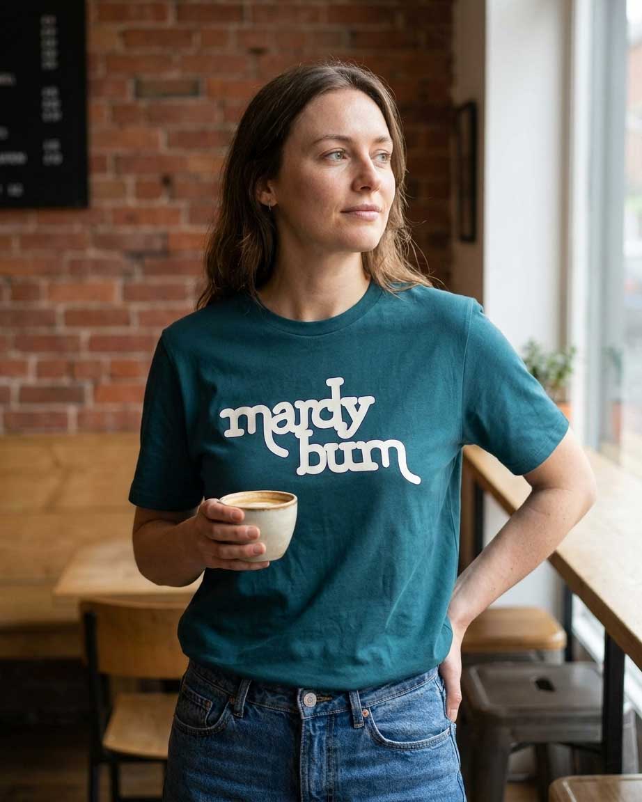 A woman wearing a teal t-shirt with the words 'mardy bum' printed in white script, holding a cup of coffee in a cafe setting with a brick wall background.