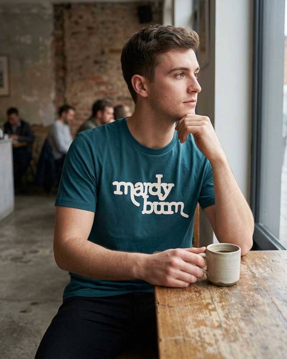A man sitting at a wooden table in a cafe, wearing a teal t-shirt with 'mardy bum' printed in white stylized text. He is holding a ceramic mug and looking out a window.