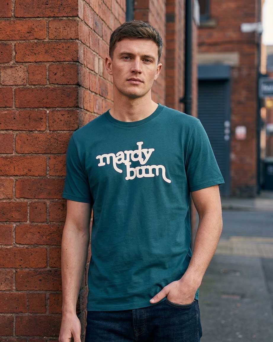 A man modeling a teal crewneck t-shirt with the phrase 'mardy bum' printed in dark blue script inspired by Arctic Monkeys across the chest, paired with denim jean against an industrial city street background.
