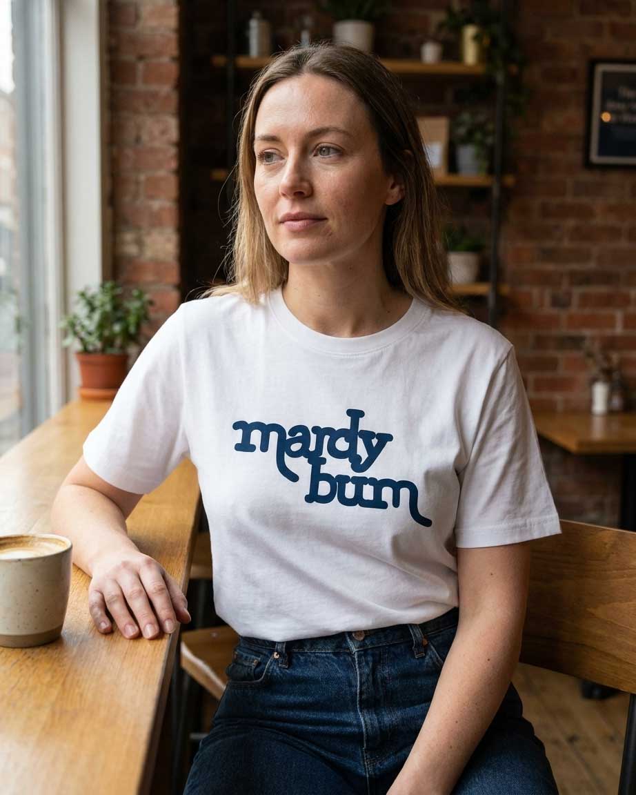 A woman sitting at a wooden table in a cozy cafe setting, wearing a white crewneck t-shirt with the phrase 'mardy bum' printed in dark blue across the chest.