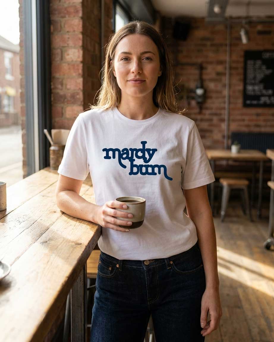 A woman standing in a rustic cafe setting wearing a white t-shirt with 'mardy bum' printed in blue text across the chest, holding a ceramic mug.