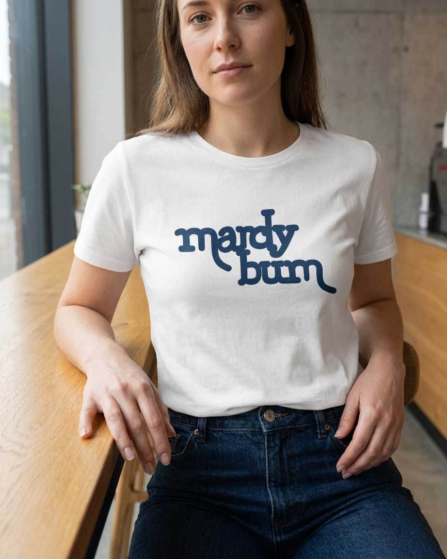 A woman wearing a white crewneck t-shirt with the phrase 'mardy bum' printed in dark blue script across the chest, paired with dark blue jeans, sitting at a wooden table.