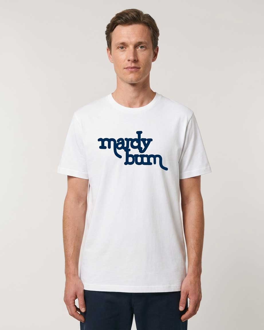 A man wearing a white crew-neck t-shirt with the words 'mardy bum' printed in dark blue across the chest, paired with dark navy pants, against a light gray studio background.