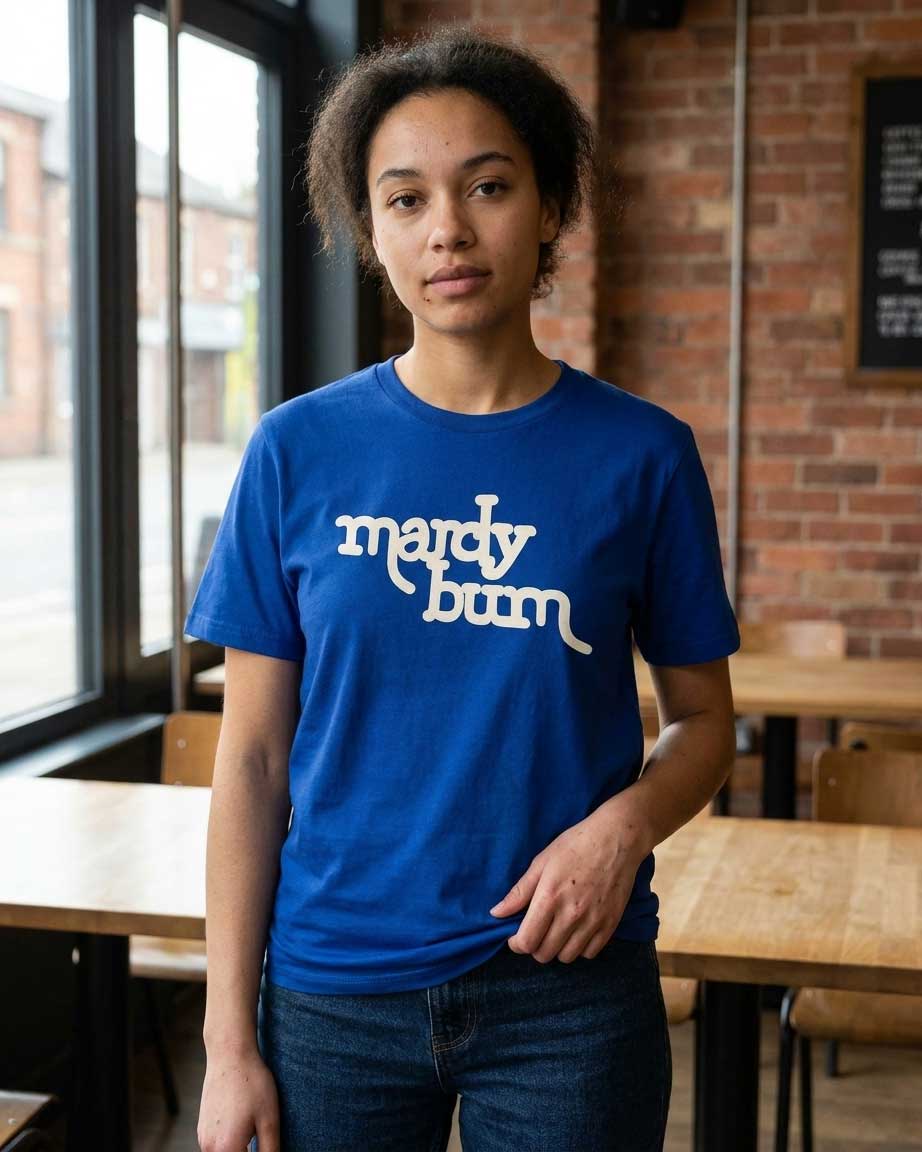 A woman wearing a royal blue crewneck t-shirt with 'mardy bum' printed in white across the chest, standing in a cafe with a brick wall and wooden tables in the background.
