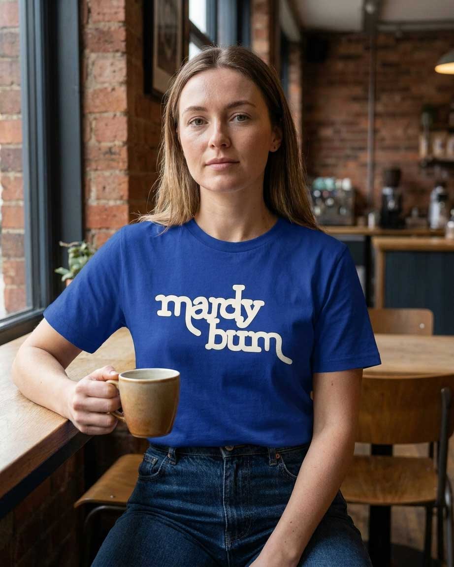 A woman sitting in a cafe wearing a royal blue t-shirt with 'mardy bum' printed in white script across the chest, holding a ceramic mug.