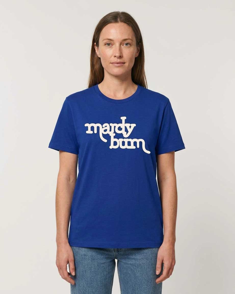 A woman wearing a royal blue short-sleeve t-shirt with 'mardy bum' printed in white across the chest, paired with blue jeans, against a plain light gray background.