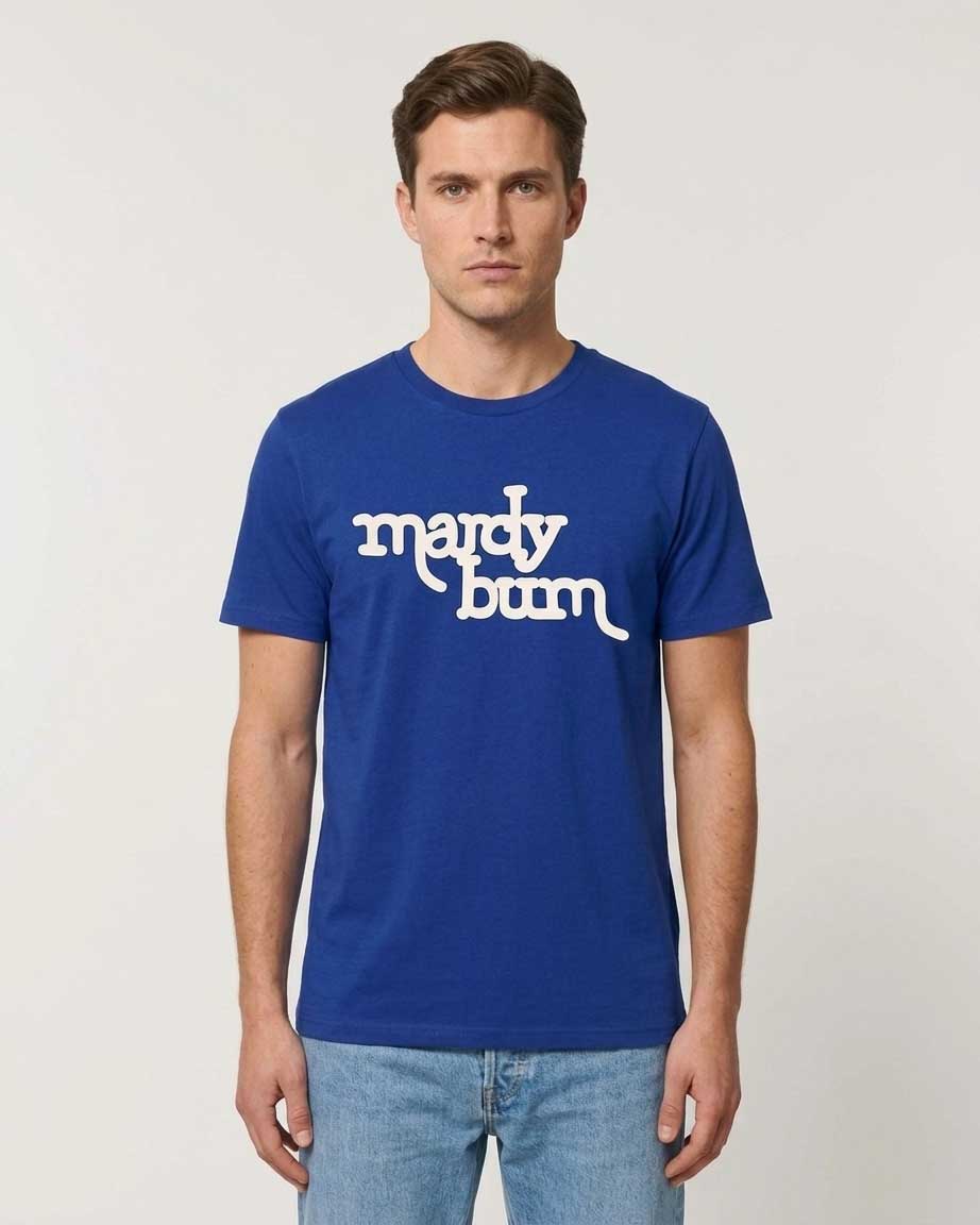 A man wearing a royal blue crewneck t-shirt with 'mardy bum' printed in white retro-style text across the chest, paired with light-wash blue jeans, against a plain light gray studio background.