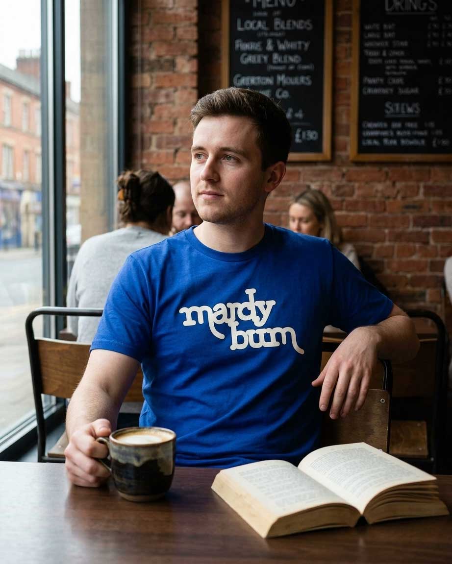 A man sitting at a cafe table wearing a blue t-shirt with 'mardy bum' printed in white. He is holding a coffee mug and an open book is on the table in front of him.