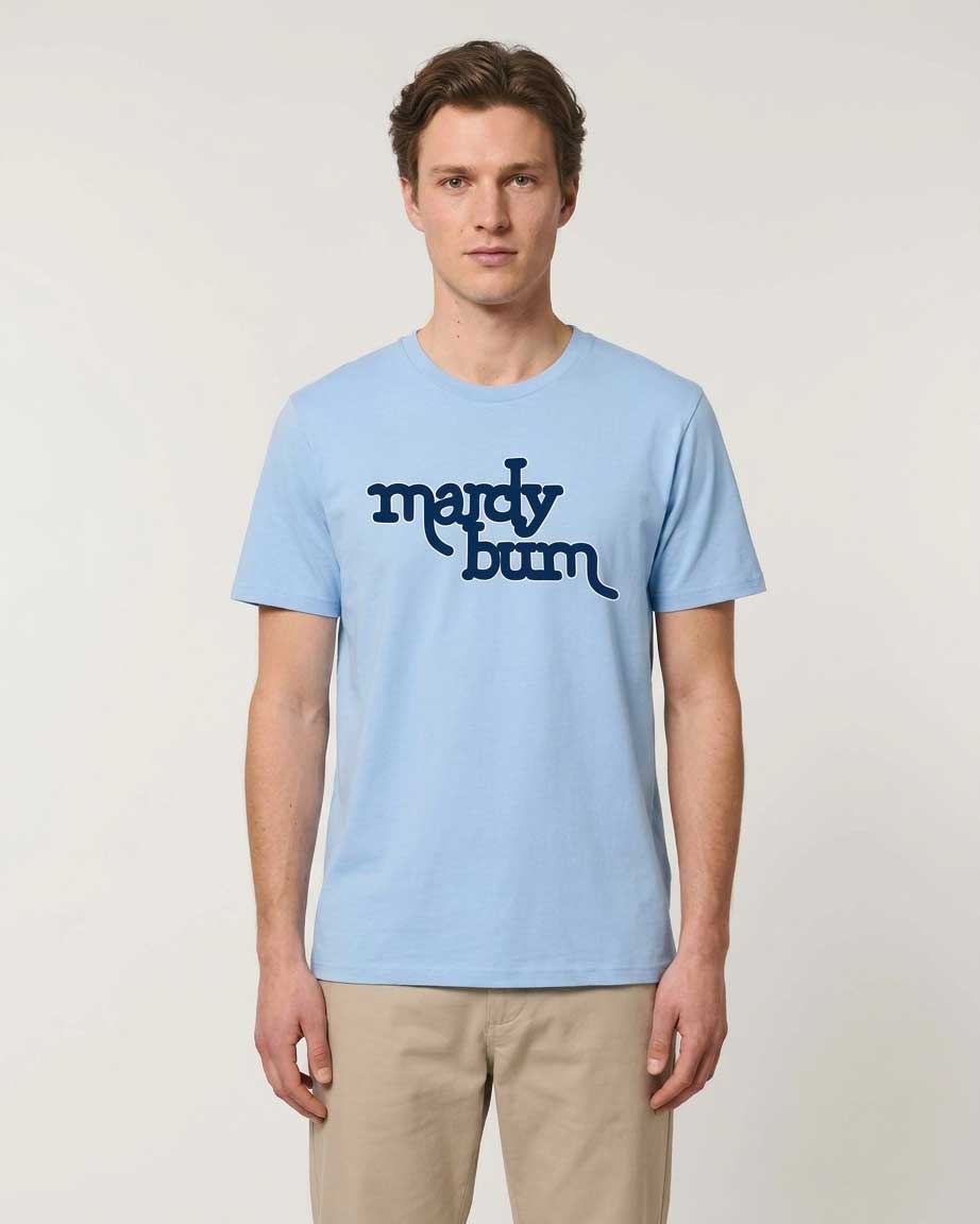 A man wearing a light blue crewneck t-shirt with the text 'mardy bum' printed in dark navy across the chest, paired with beige trousers, against a plain light gray background.
