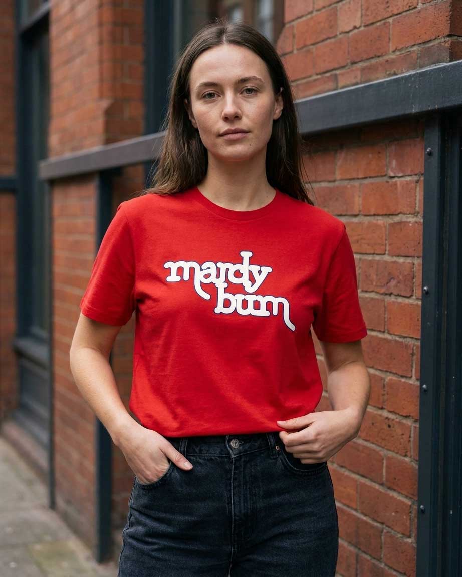 A woman wearing a bright red crewneck t-shirt with the words 'mardy bum' printed in white stylized text across the chest, paired with dark blue jeans, standing against a red brick wall.
