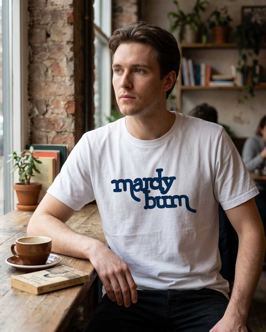 A man sitting at a wooden table in a cozy cafe setting, wearing a white crewneck t-shirt with the words 'mardy bum' printed in dark blue script across the chest.