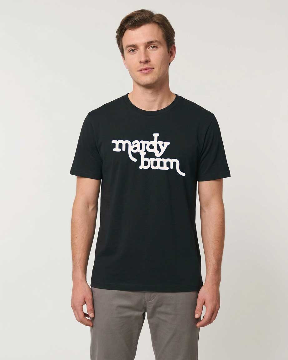 A man wearing a black crewneck t-shirt with 'mardy bum' printed in white script across the chest, paired with gray pants, standing against a light gray studio background.