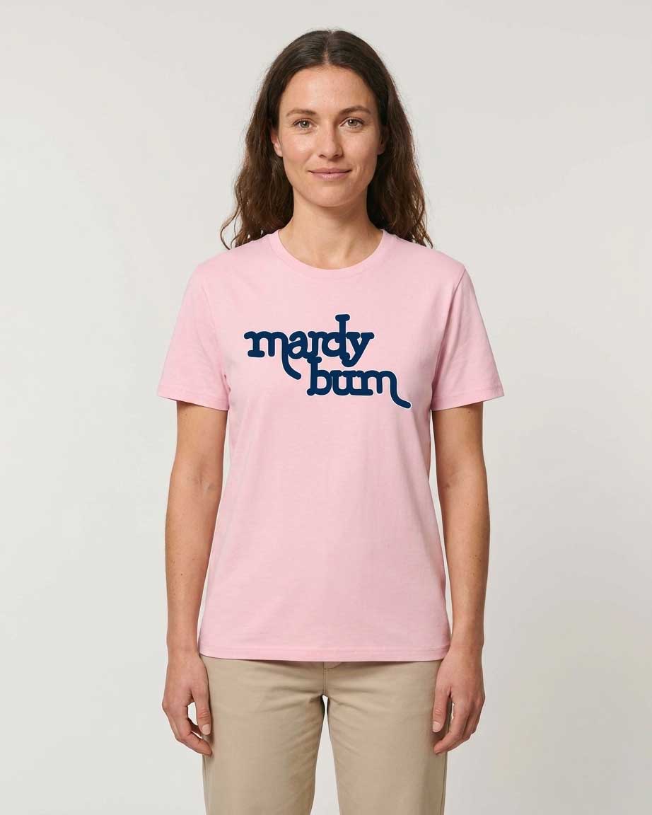 A woman modeling a light pink crewneck t-shirt with the phrase 'mardy bum' printed in dark blue script inspired by Arctic Monkeys across the chest, paired with beige pants, against a plain light gray background.