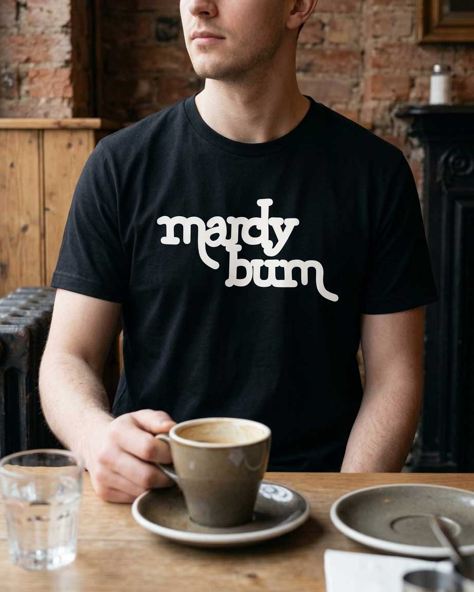 A man wearing a black t-shirt with the phrase 'mardy bum' printed in white stylized text, sitting at a wooden table with a coffee cup in a cafe setting.